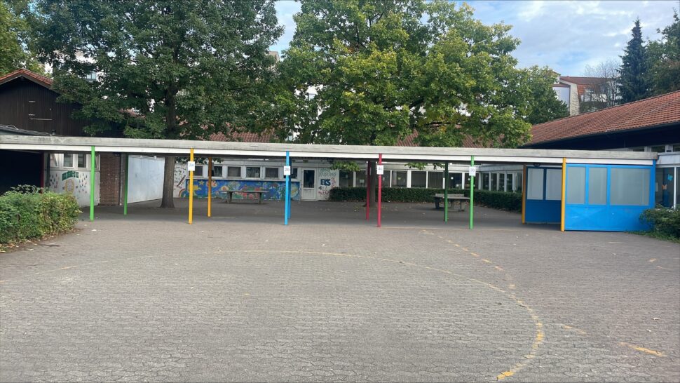 Schulhof mit einem überdachten Gang, der von verschiedenfarbig gestrichenen Pfosten getragen wird, umgeben von Gebäuden und Bäumen im Hintergrund.
