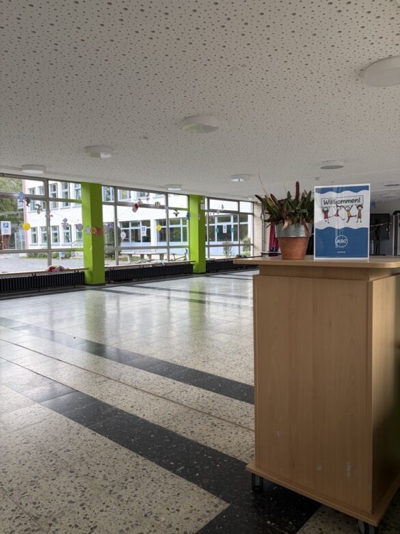 Ein Schulflur mit grünen Säulen, großen Fenstern, Fliesenboden und einem Holzschrank, in dem eine Topfpflanze und ein Schild mit der Aufschrift „Willkommen!“ stehen.